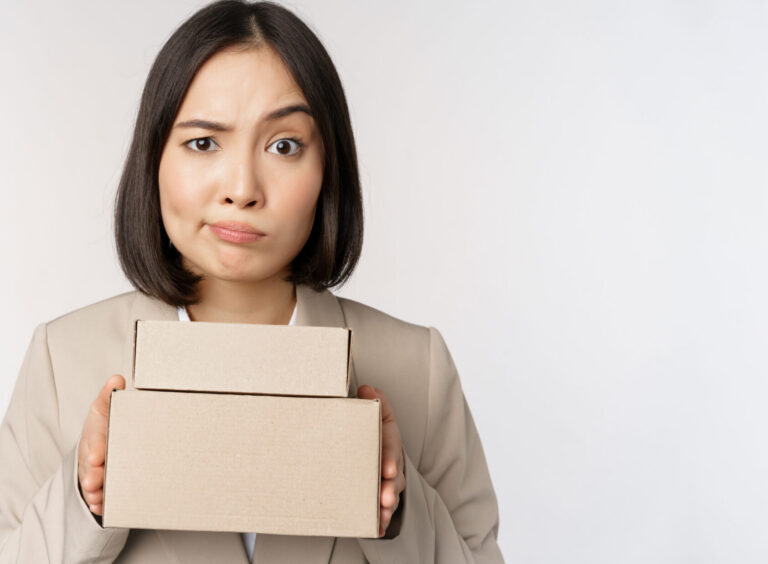 Portrait of asian saleswoman, female entrepreneur holding boxes and looking sad, disappointed, standing over white background