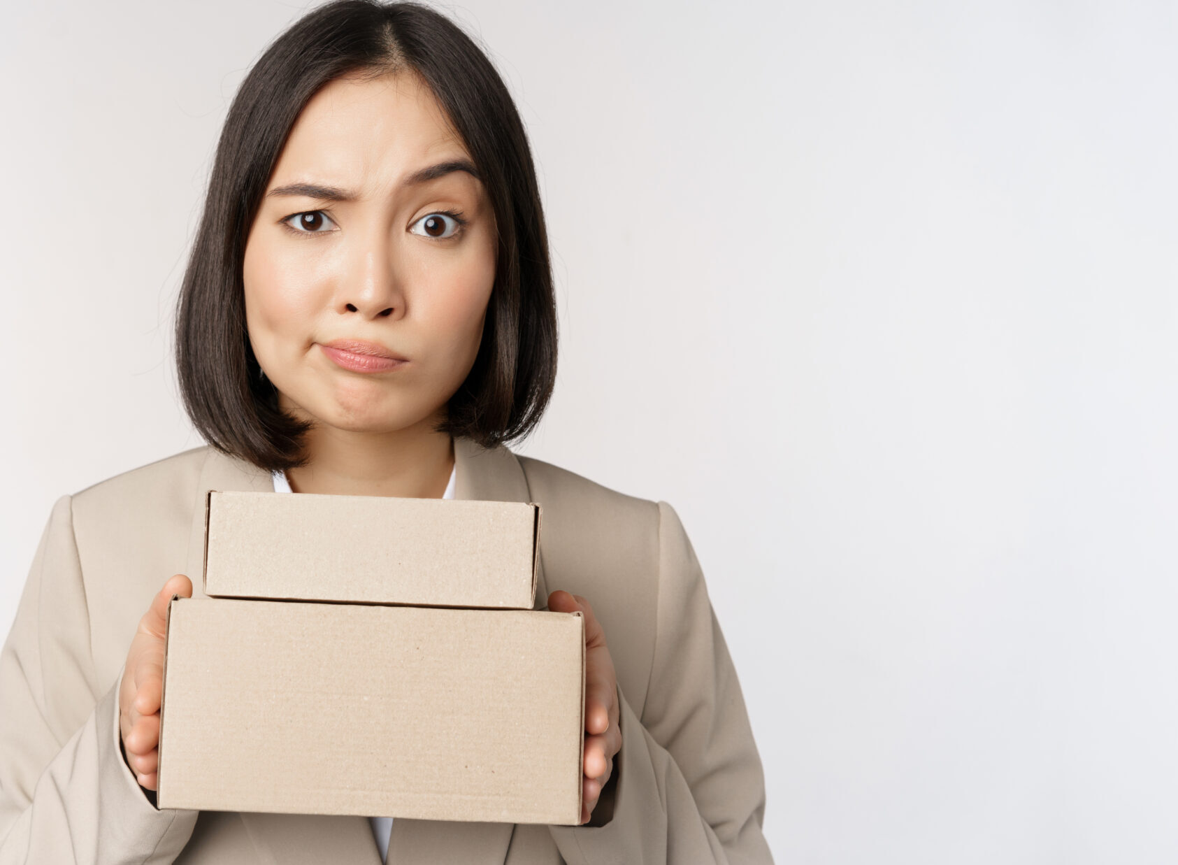 Portrait of asian saleswoman, female entrepreneur holding boxes and looking sad, disappointed, standing over white background